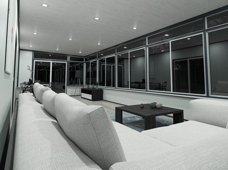 Black and white photo of the interior of a sunroom with windows along all of the walls except where there is a door with windows, a solid roof, and a long couch along the wall in the foreground.